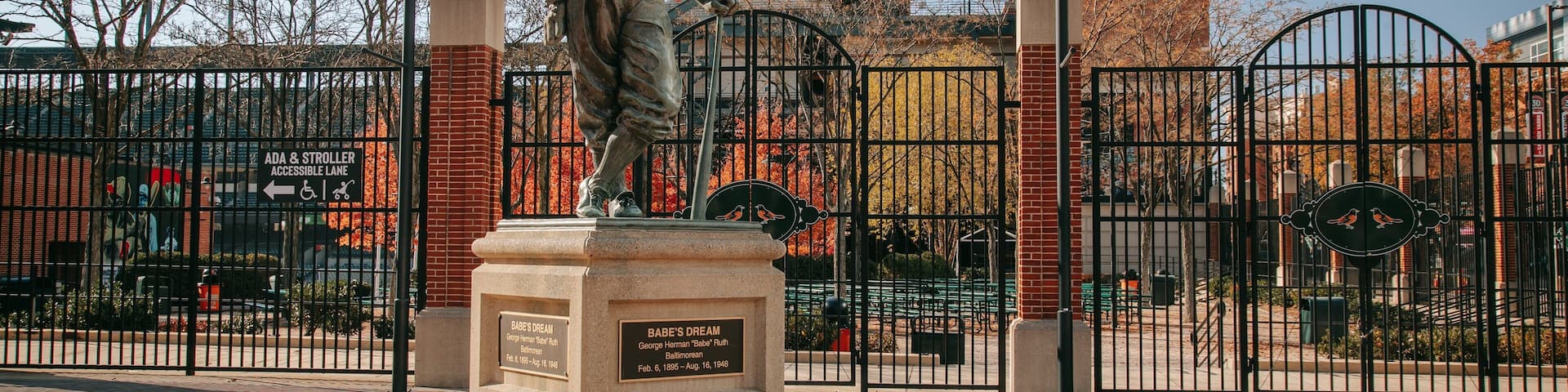Una estatua de Babe Ruth en un parque, con un muro de ladrillo y rejas metálicas al fondo.
