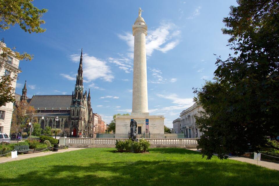 Un monumento alto y blanco con inscripciones, rodeado de un césped cuidado y edificios históricos.