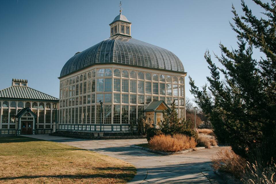 Un invernadero grande con cúpula de vidrio y una estructura más pequeña adosada, rodeado de césped y árboles.