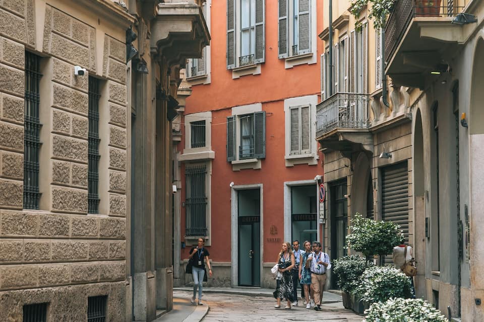 Eine schmale Gasse mit Steinhäusern, ein rotes Gebäude mit Fensterläden und Passanten.