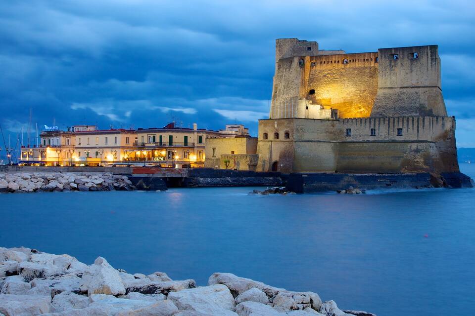 A historic castle by a calm sea, with a well-lit waterfront and boats in the background.