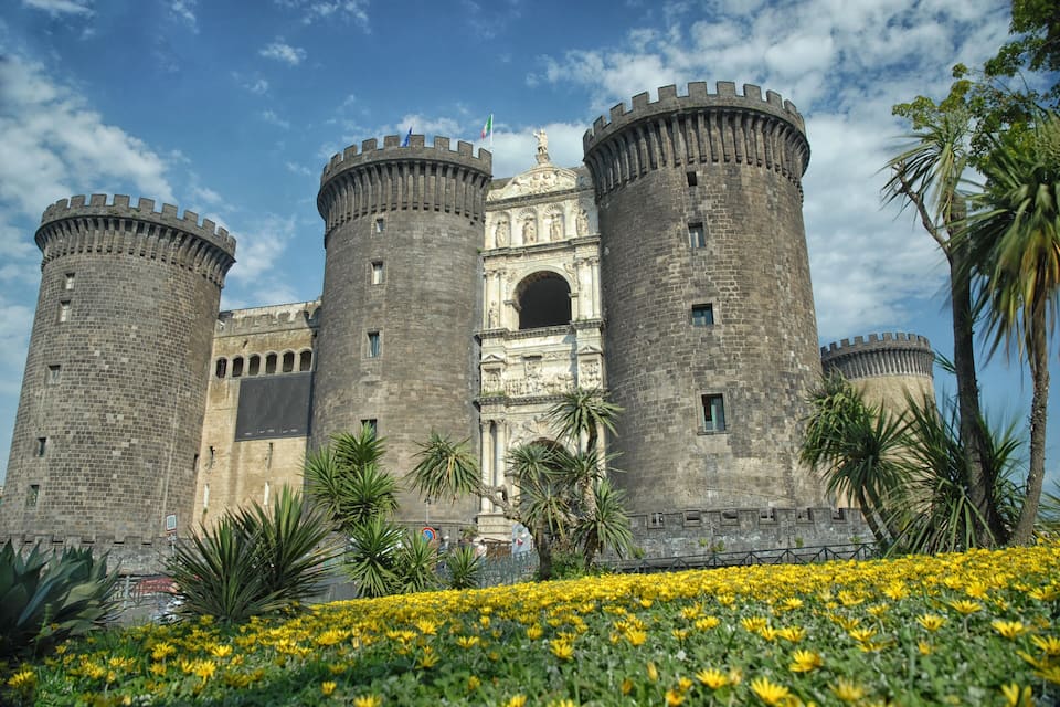 A historic castle with four towers, a central entrance, and a surrounding garden with yellow flowers.