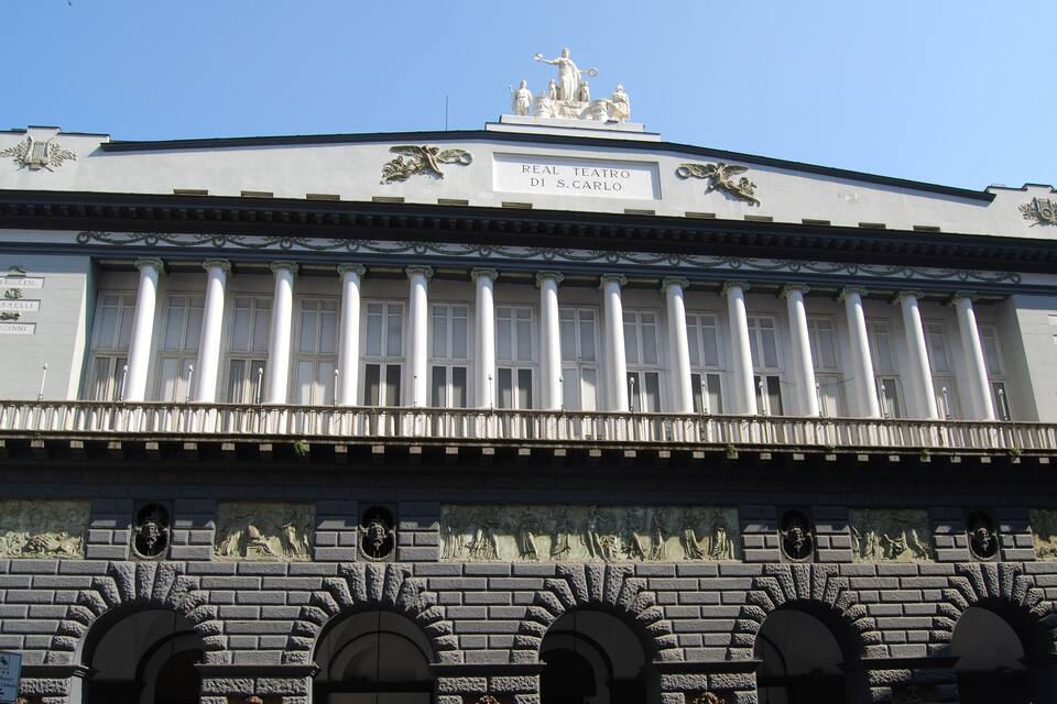 A classical building with columns, a statue on top, and a plaque reading "Real Teatro Di S. Carlo".