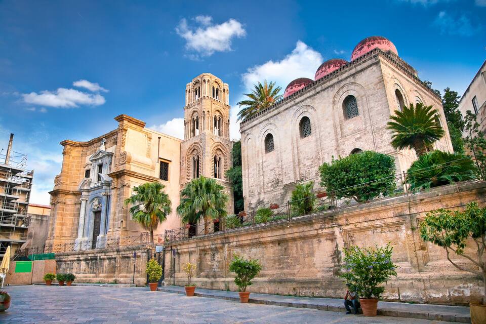 Un edificio histórico con un campanario y ventanas arqueadas, rodeado de palmeras y plantas en macetas.