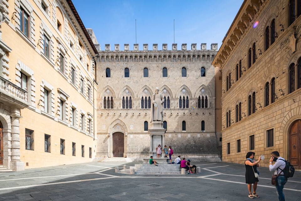 A historic courtyard with a central statue, flanked by ornate buildings with arched windows and stone facades.