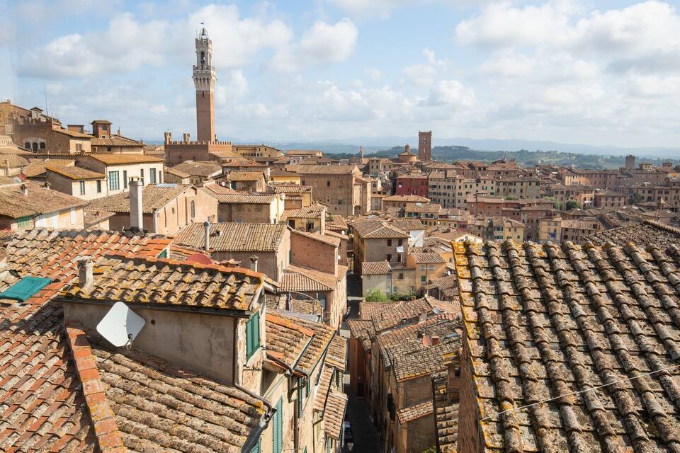 A historic town with terracotta rooftops, a prominent tower, and a mountainous backdrop.