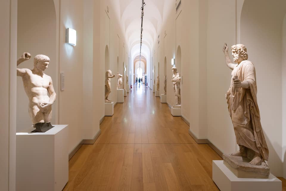 A museum corridor with marble statues on pedestals, wooden floors, and white walls.