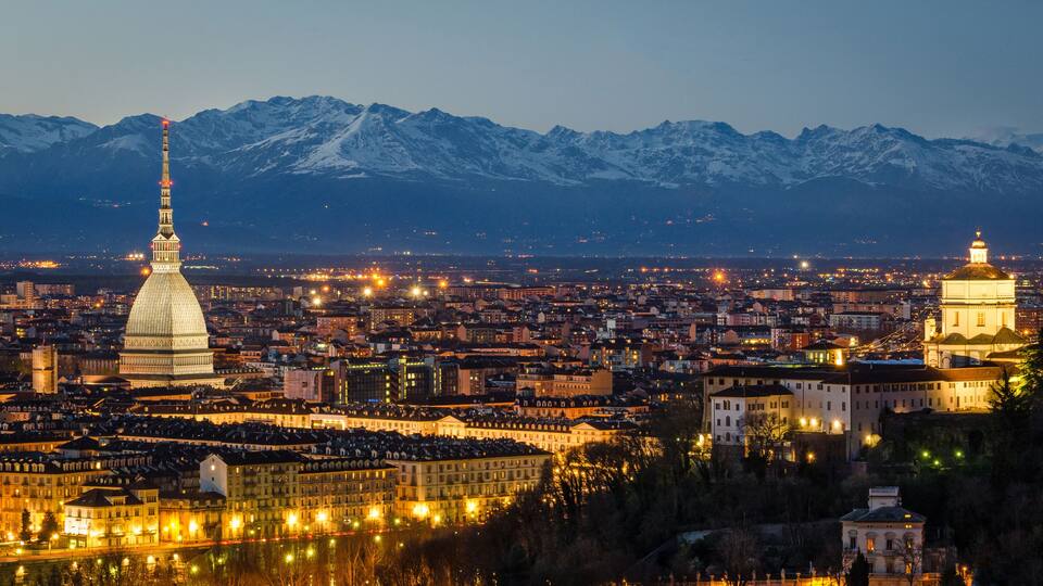 Un paisaje urbano con una cúpula destacada y una torre, con montañas nevadas al fondo.