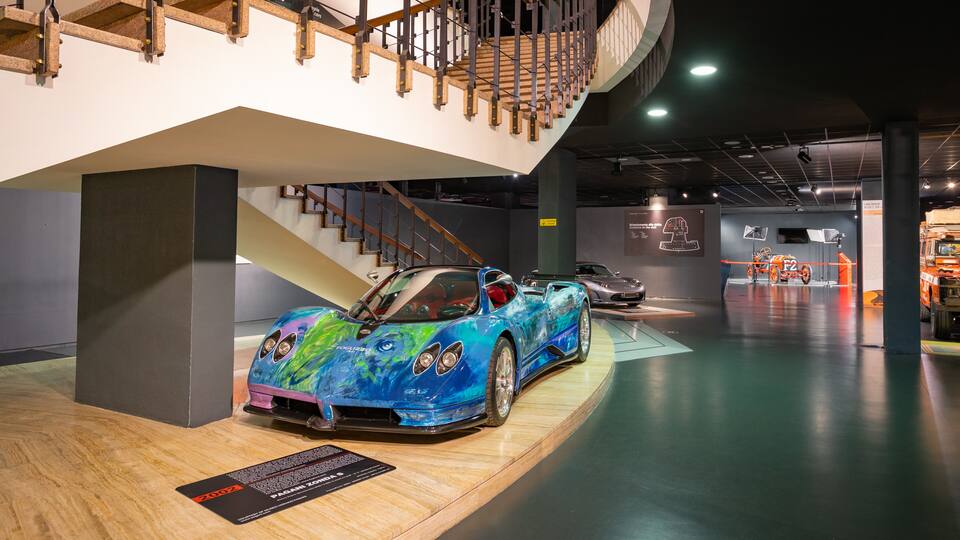 Una galería con un coche deportivo en exhibición, escaleras de madera y un camión al fondo.