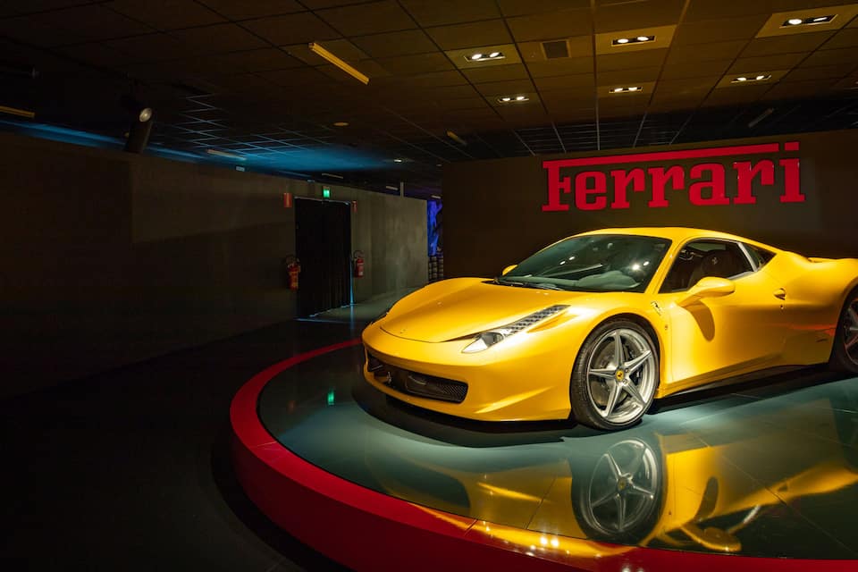 Un cotxe esportiu Ferrari groc exposat en un taller, amb un disseny de sòl vermell i negre.