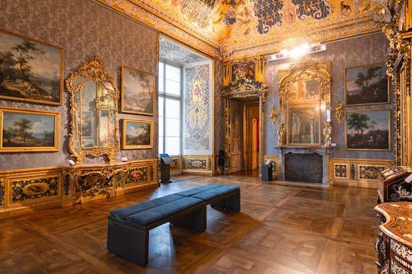 Una habitación opulenta con pinturas enmarcadas en oro ornamentado, una chimenea imponente y un amplio banco.