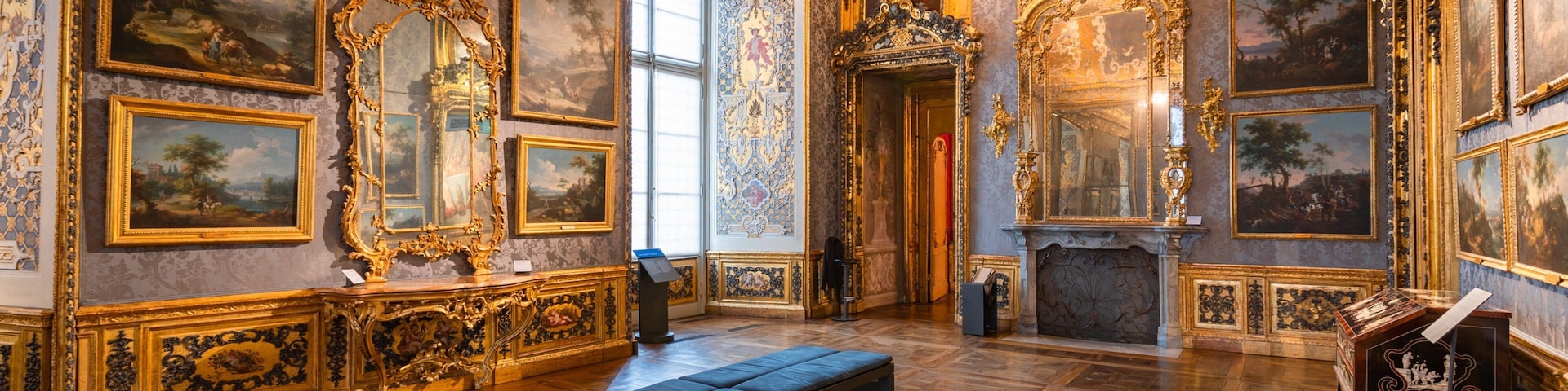 Um quarto luxuoso com pinturas emolduradas em ouro trabalhado, uma lareira imponente e um amplo banco.
