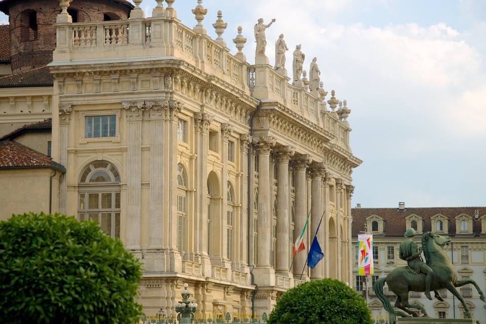 Um edifício histórico imponente com colunas e estátuas ornamentadas, incluindo uma estátua de um cavaleiro montado e bandeiras.
