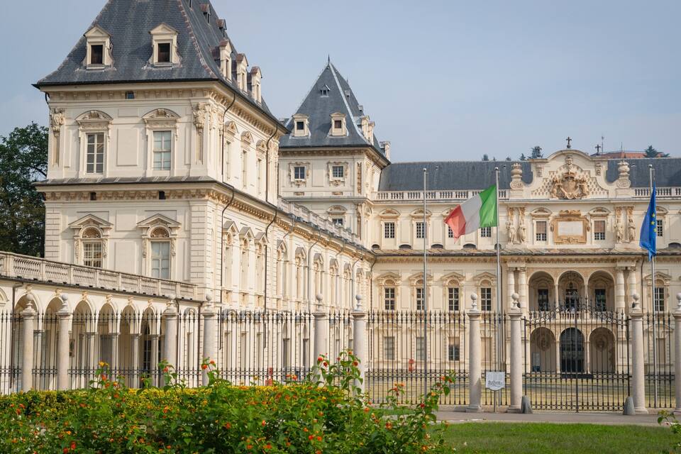 A grand historic building with ornate architecture, featuring multiple flags, including the Italian flag, and a well-maintained garden with vibrant flowers.