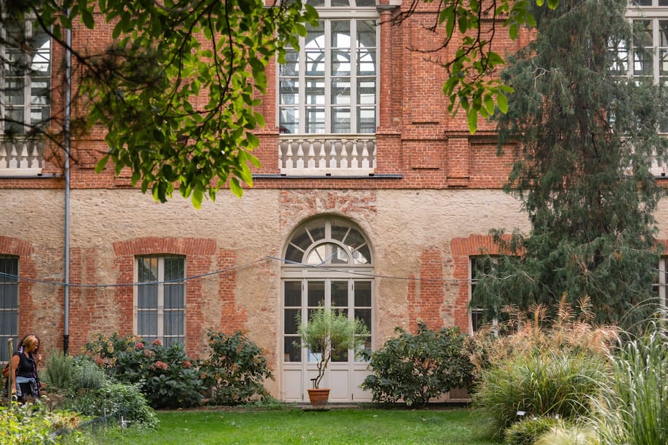 A red brick building with arched windows and a white door, surrounded by a well-maintained garden with various plants and a lawn.