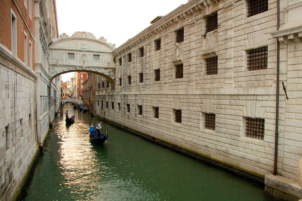 Un canal avec une gondole, un bâtiment en pierre doté de fenêtres en arc de cercle et un pont en arrière-plan.