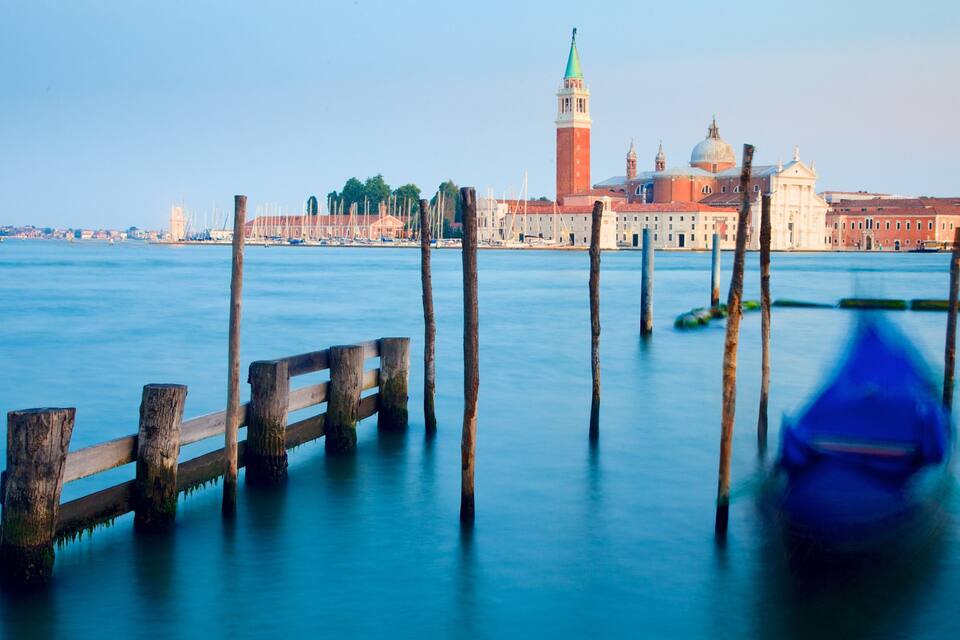 A waterfront with a wooden pier, a gondola, and historic buildings in the background.
