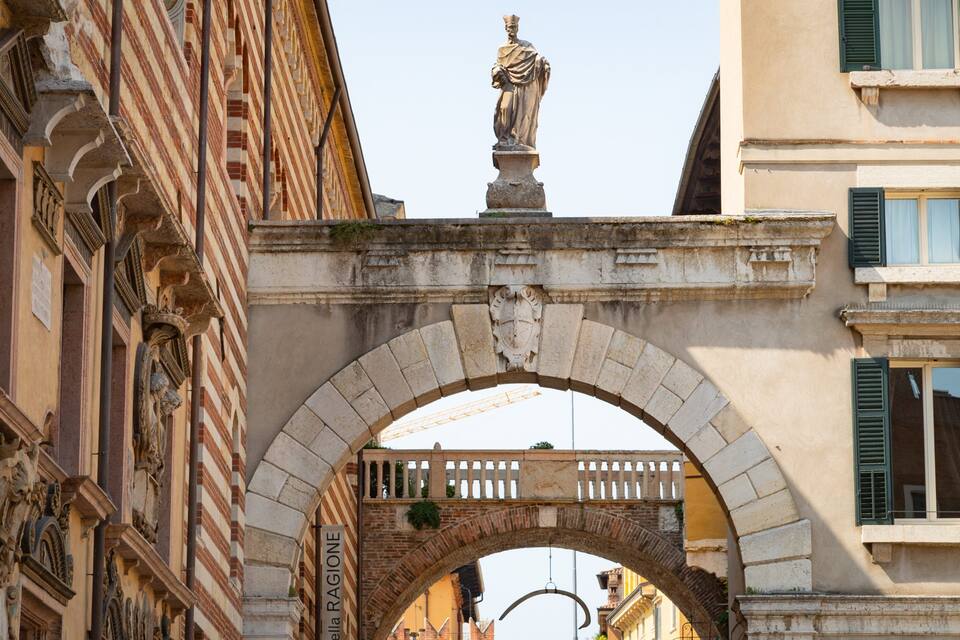 Un puente de piedra arqueado con una estatua en la parte superior, rodeado de edificios históricos con fachadas ornamentadas y contraventanas verdes.