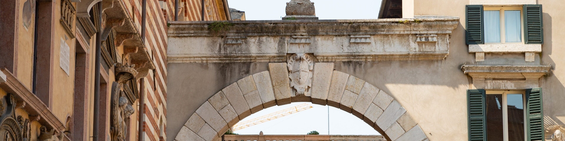 Uma ponte de pedra arqueada com uma estátua no topo, rodeada por edifícios históricos com fachadas ornamentadas e persianas verdes.