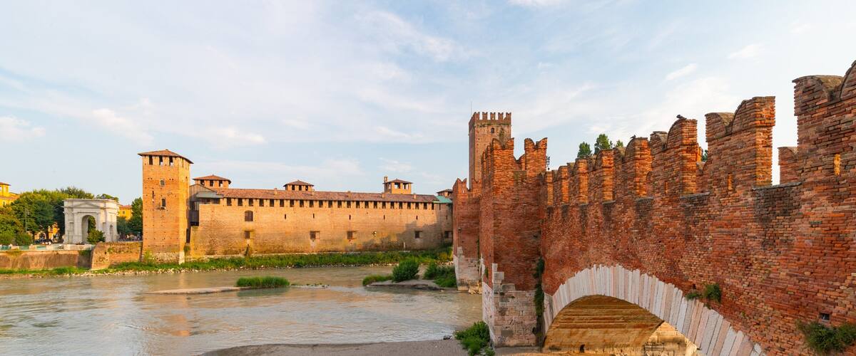 A historic brick bridge over a river, with a fortified wall and a tower in the background.