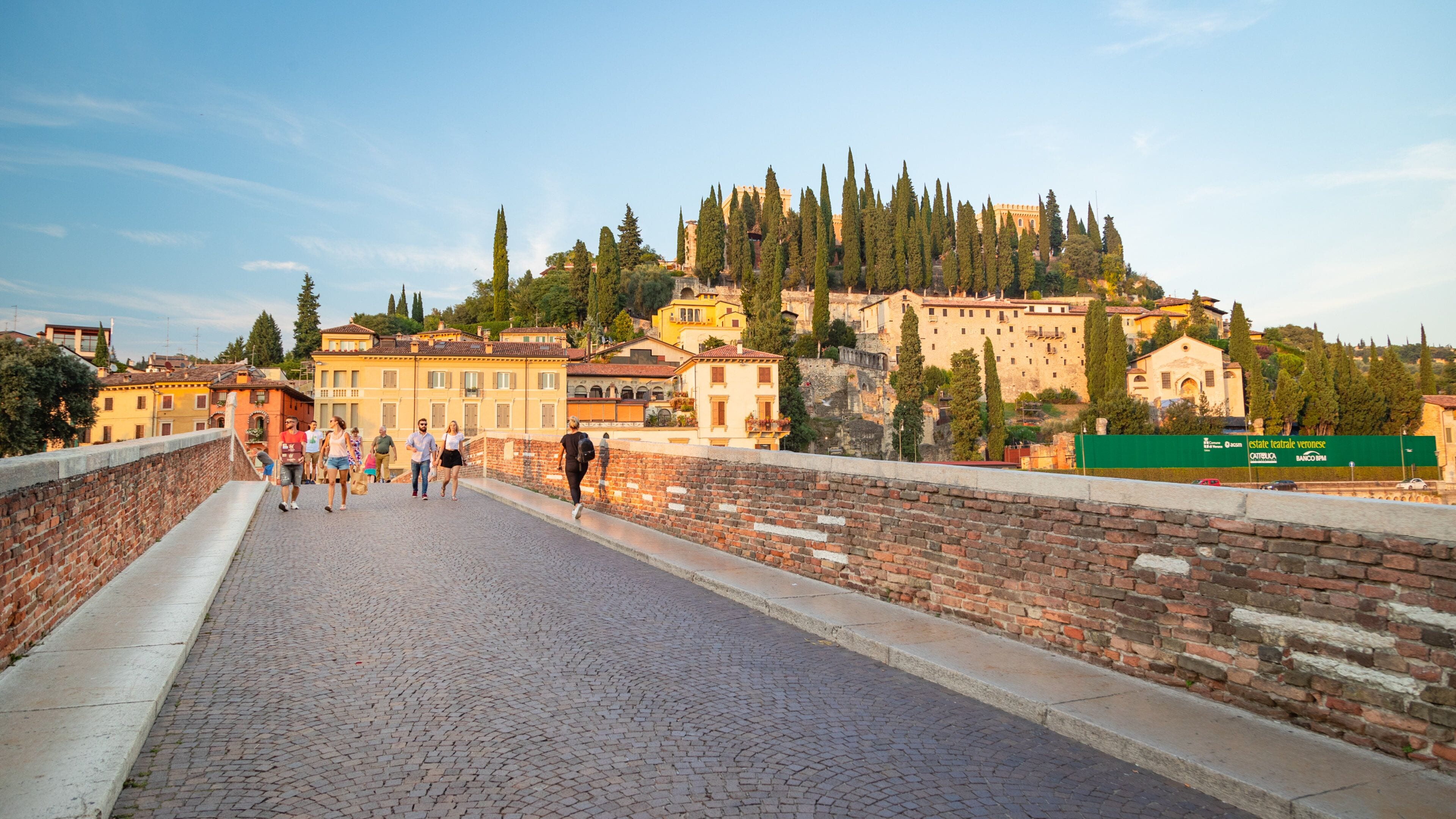 A cobblestone bridge with people walking, overlooking a historic town with buildings and trees.