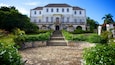 A large colonial-style building with a central staircase leading to a door, surrounded by lush gardens and palm trees.