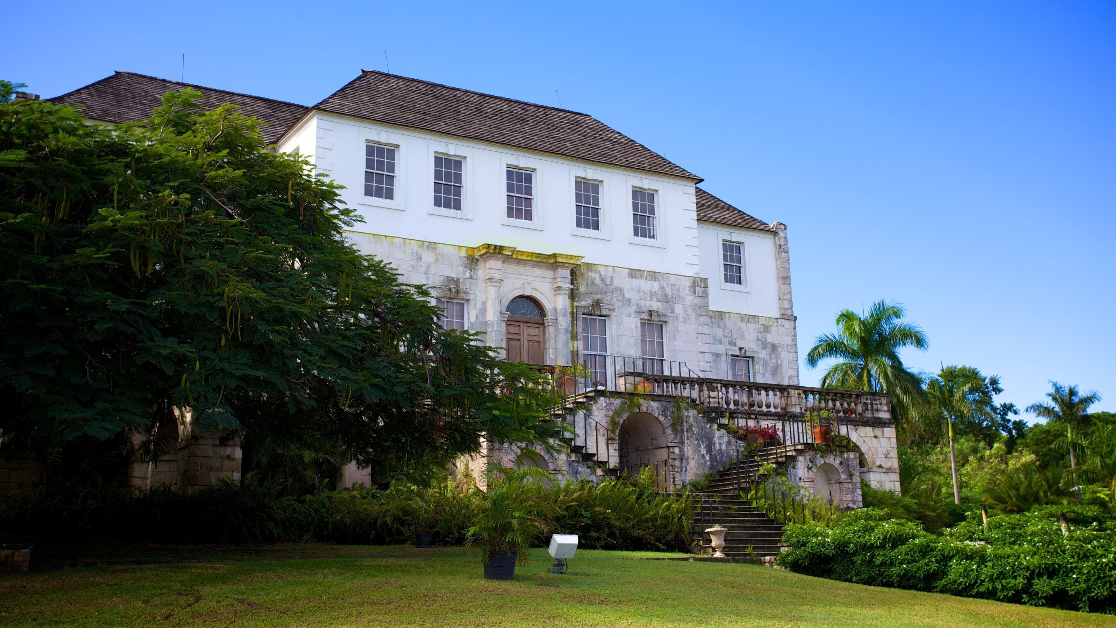A historic building with a white facade and a tiled roof, surrounded by lush greenery and palm trees.