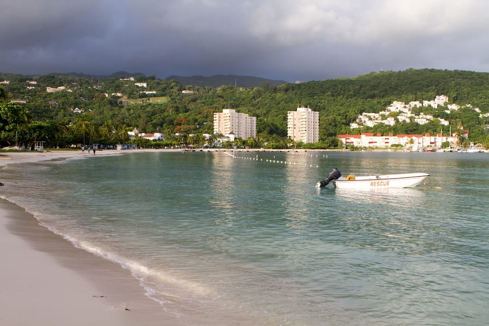 A beach with a boat named 'Rescue' anchored in calm waters, surrounded by lush green hills and buildings in the background.
