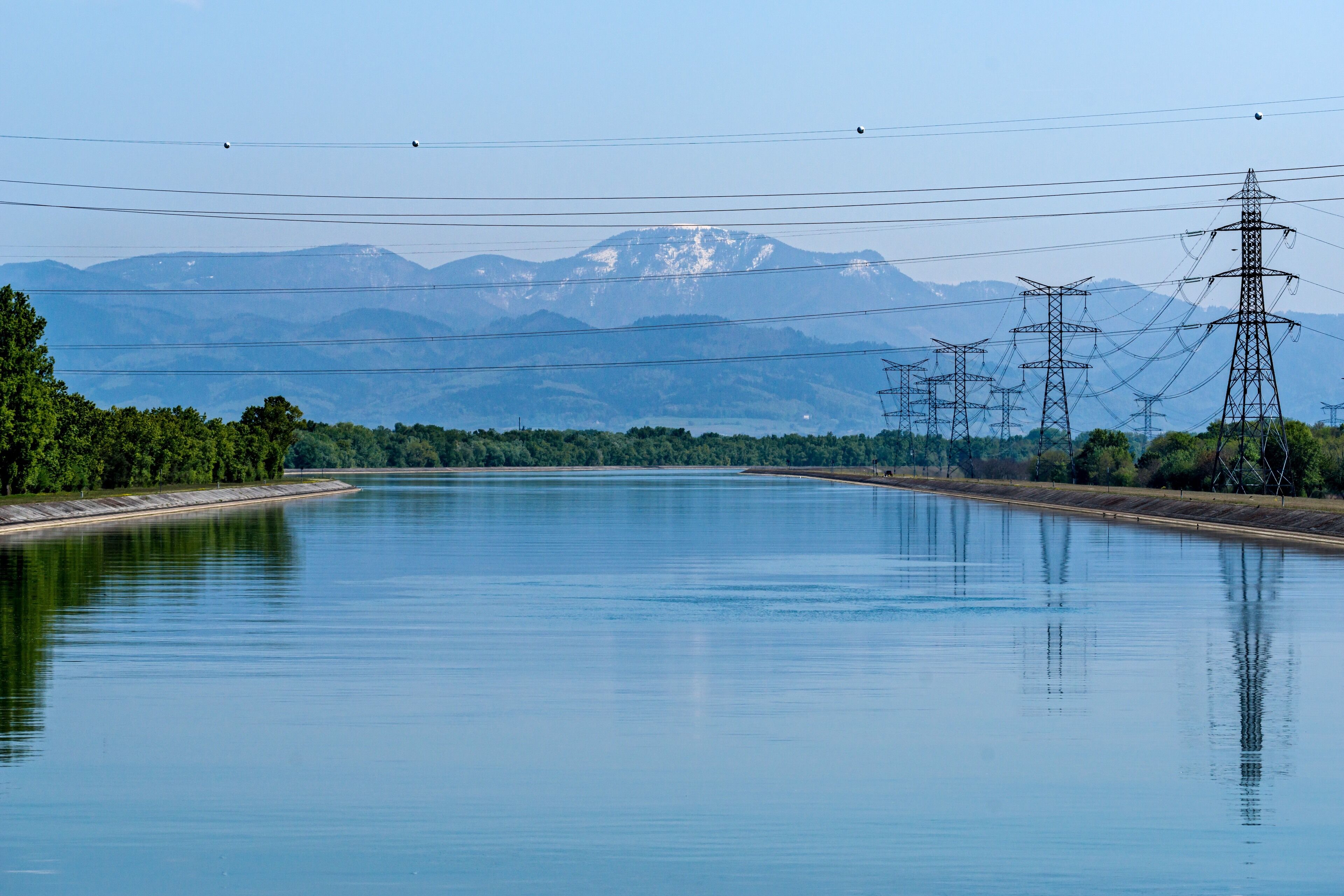 A calm river with mountains in the background and power lines running parallel to the water.