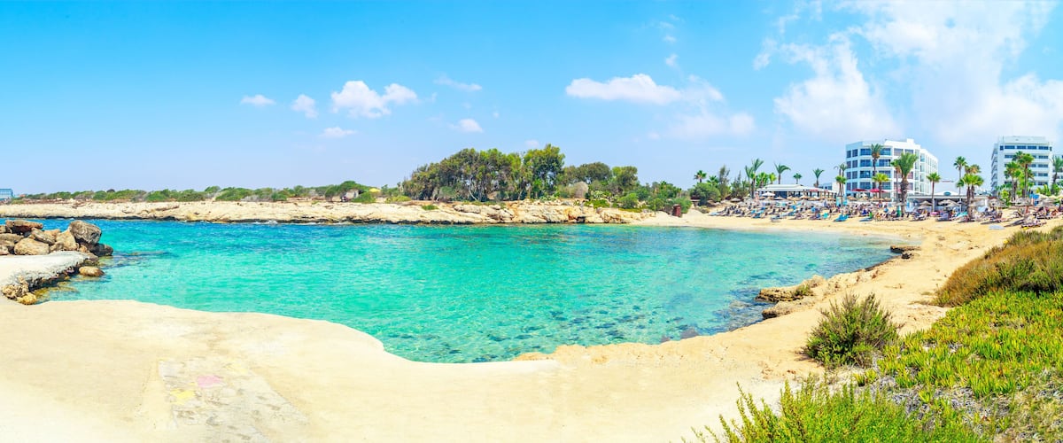 A beach with clear turquoise water, sandy shore, rocky outcrops, and palm trees.