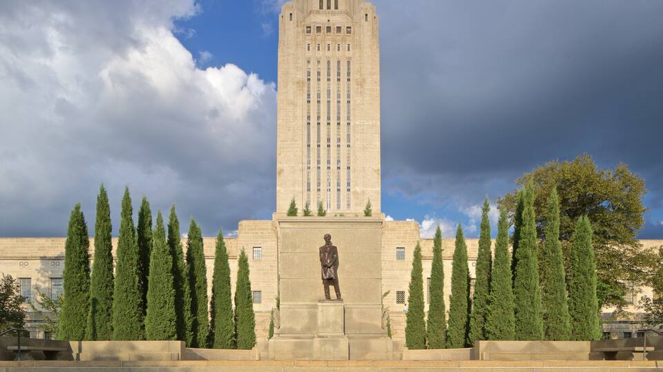 A tall, beige stone monument with a statue in front, surrounded by rows of tall, green cypress trees.