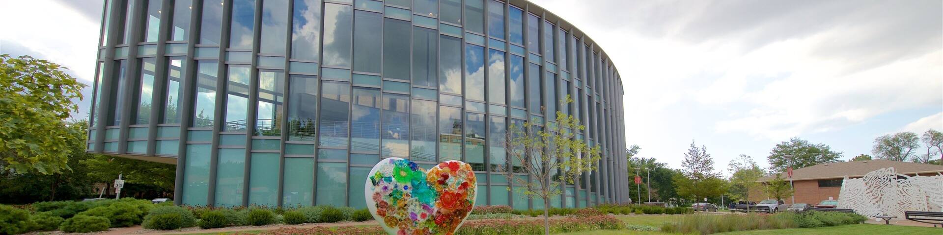 Un edificio moderno con fachada de vidrio, una escultura con forma de corazón en un área ajardinada y un árbol en primer plano.
