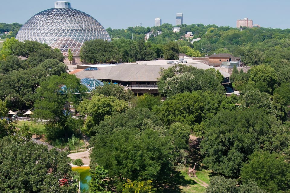 Uma grande cúpula geodésica, um jardim zoológico com girafas, e uma paisagem urbana ao fundo.