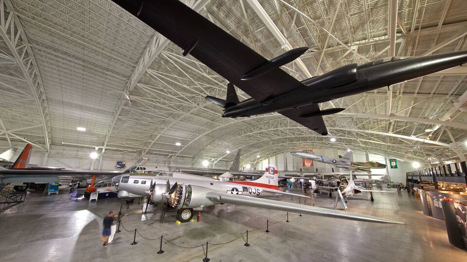 A large indoor hangar with various aircraft on display, including a prominent fighter jet and a vintage propeller plane.