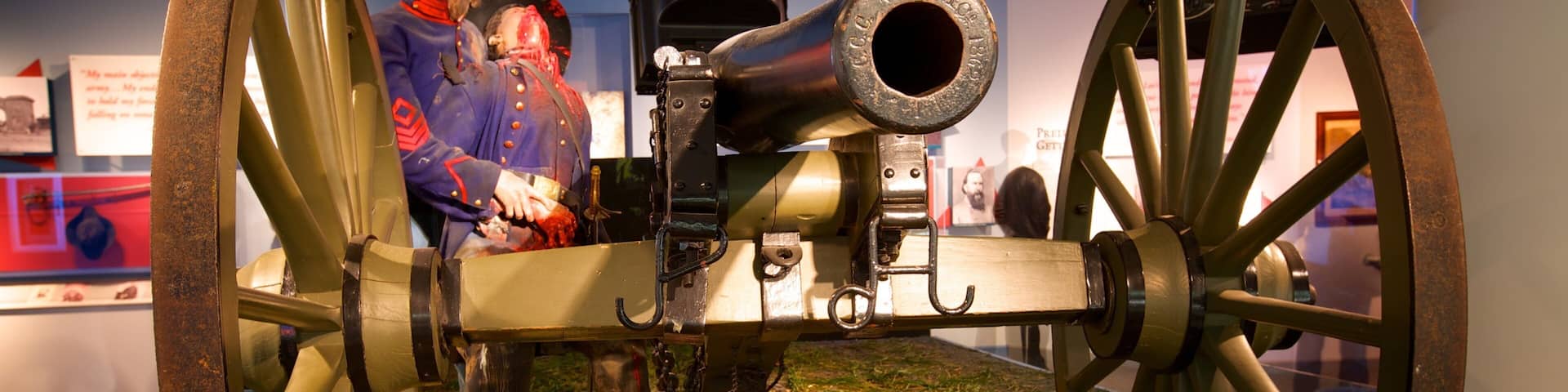 A historical cannon with large wheels, mounted on a grassy display with two figures in period uniforms.