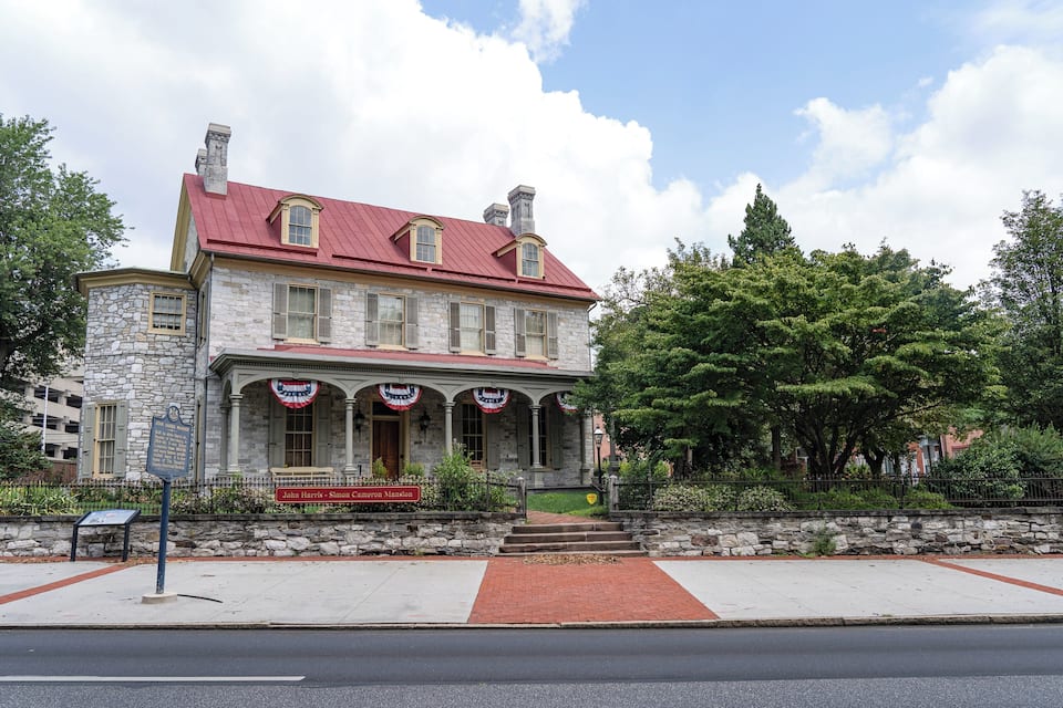 A historic stone building with a red roof and a sign reading "John Harrelson - Simon Cameron Mansion".