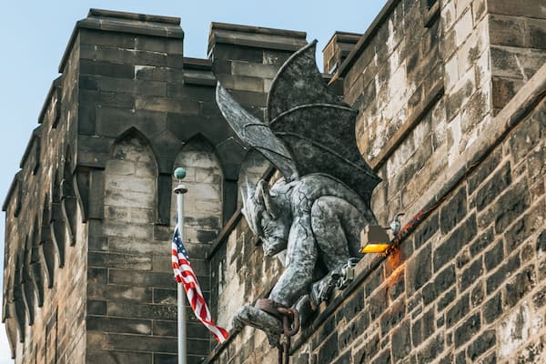 A gargoyle perched on a stone wall with an American flag in front.