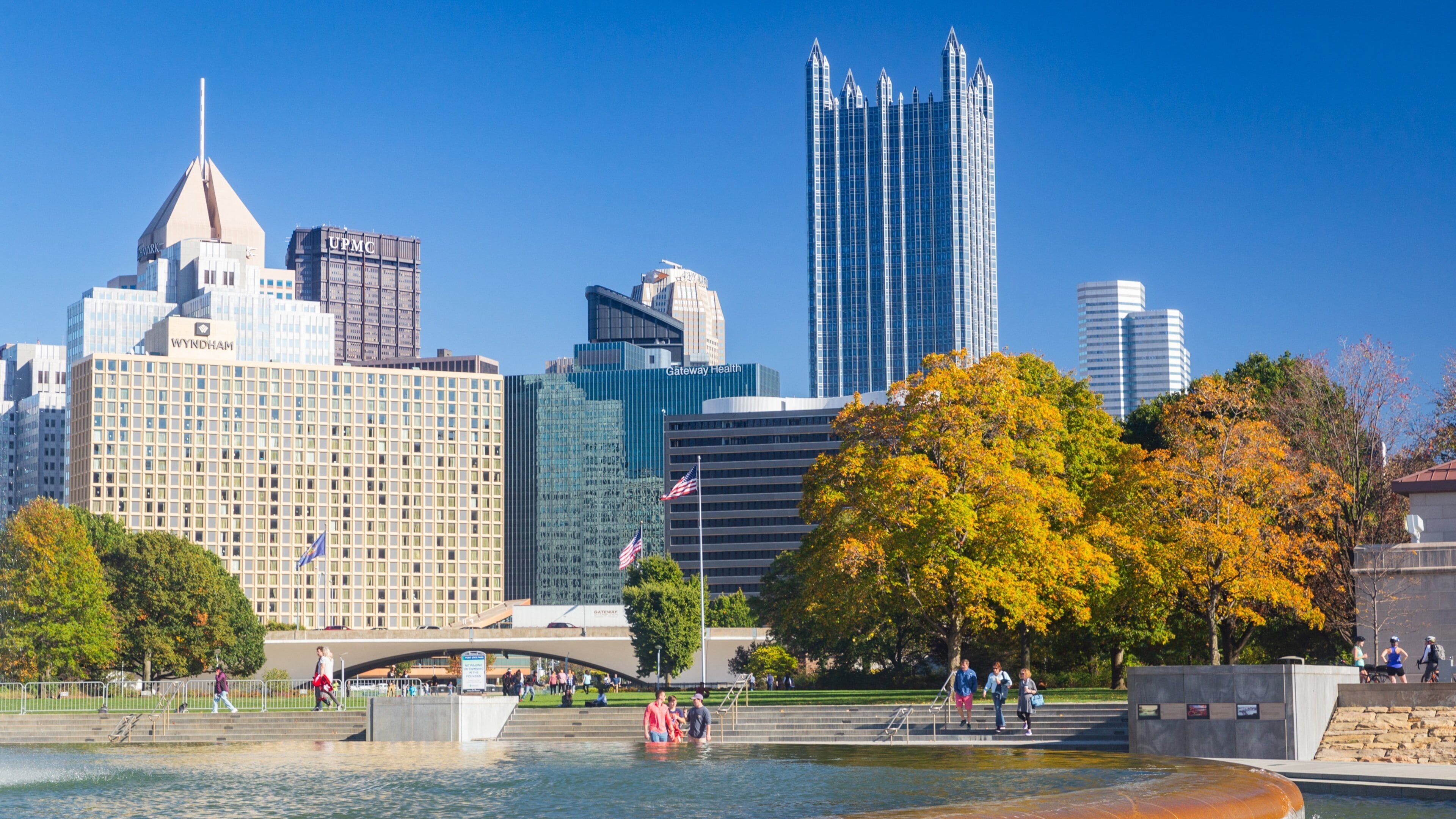 Linha do horizonte urbano com edifícios notáveis como a UPMC e Gateway Health, uma orla marítima com pessoas caminhando e árvores com folhagem avermelhada outonal.