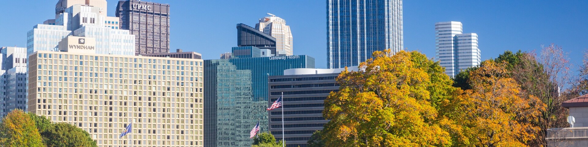 Linha do horizonte urbano com edifícios notáveis como a UPMC e Gateway Health, uma orla marítima com pessoas caminhando e árvores com folhagem avermelhada outonal.
