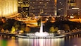 Panoramica notturna di una città con una fontana centrale, edifici illuminati e un ponte sullo sfondo.