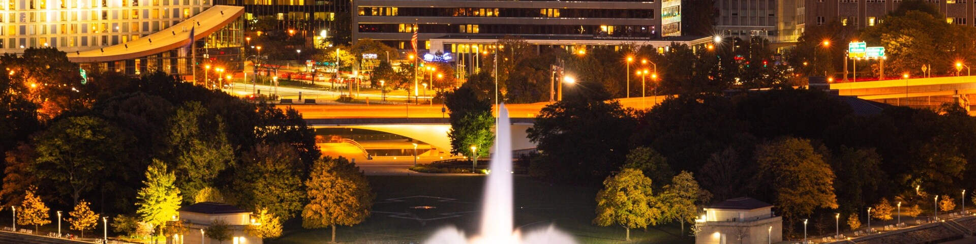 A cityscape at night with a central fountain, illuminated buildings, and a bridge in the background.