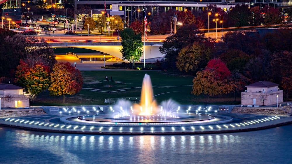 A fountain in a park with autumn trees and a modern building in the background.