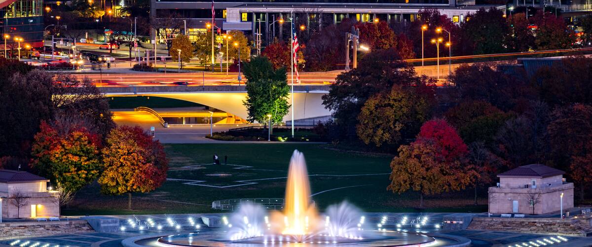 A fountain in a park with autumn trees and a modern building in the background.