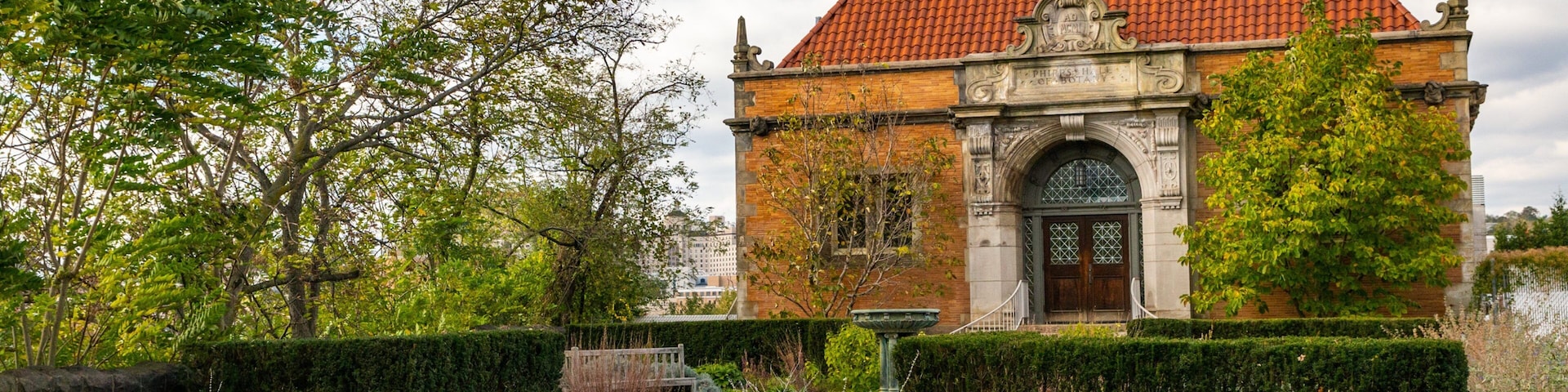 Um edifício histórico com telhado de telhas vermelhas, rodeado por jardins e árvores bem cuidados.