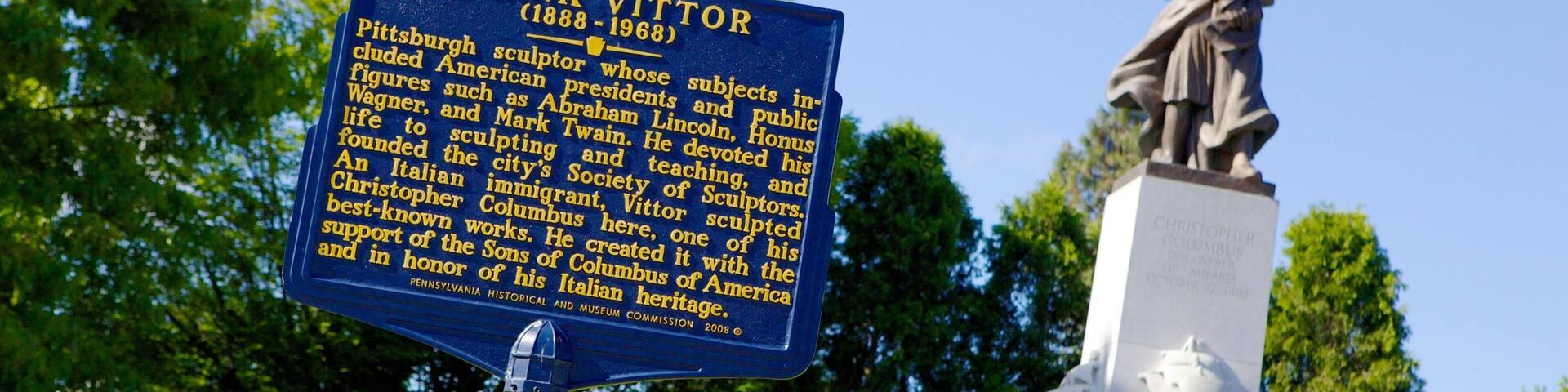 A blue historical marker with gold lettering and a statue of Christopher Columbus in the background.
