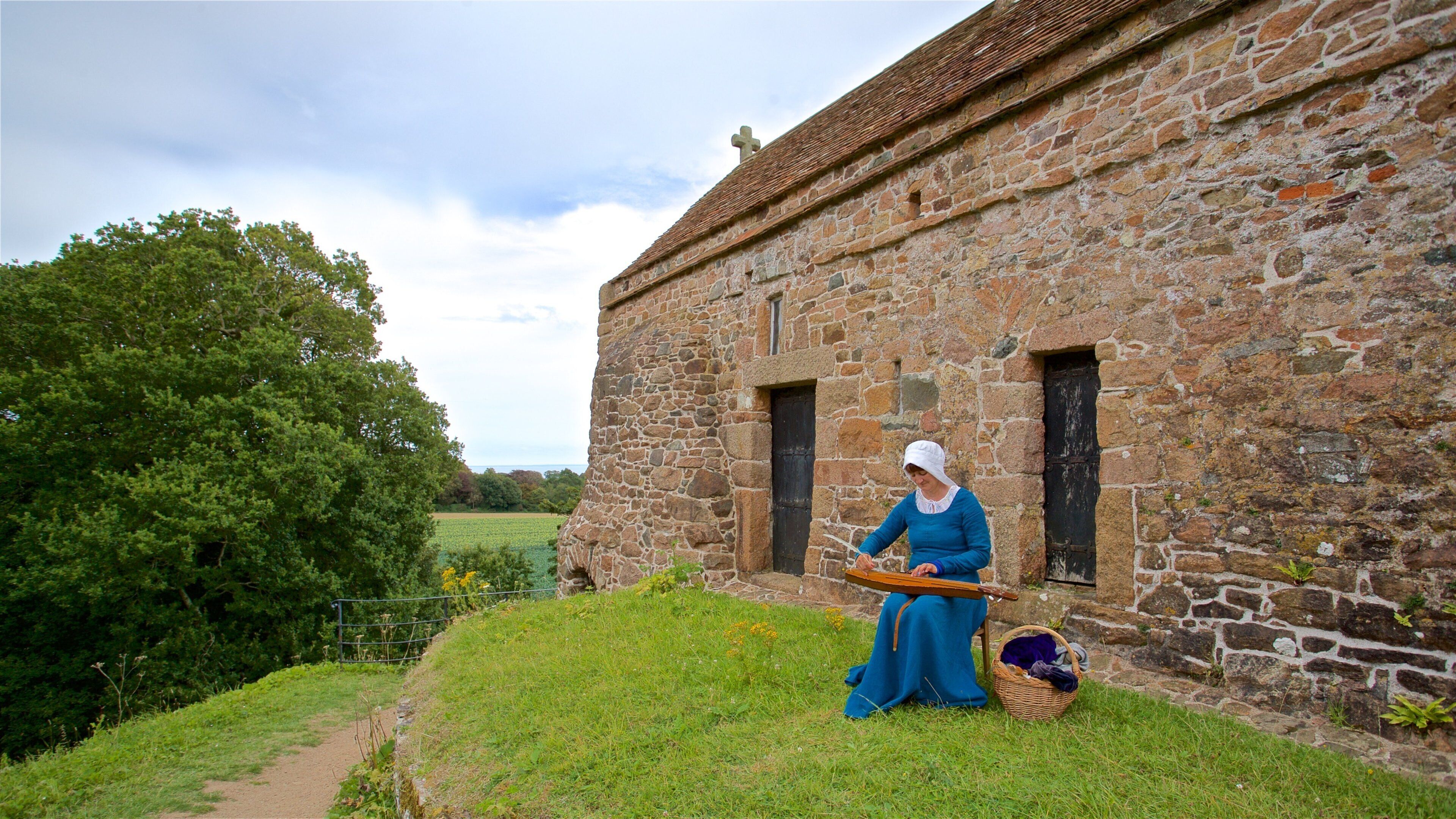 A woman in historical attire sitting on a grassy area in front of a stone building with a cross on the roof.