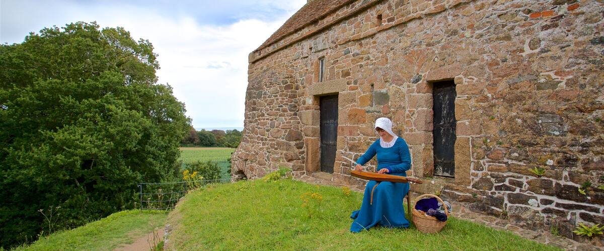 A woman in historical attire sitting on a grassy area in front of a stone building with a cross on the roof.