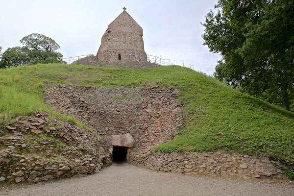 Ein steinerner Turm mit Kruzifix, umgeben von grünen Hügeln und Bäumen.