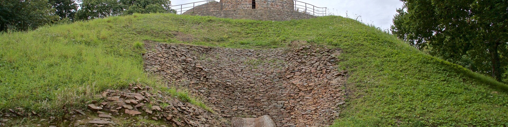 Una torre de piedra con una cruz en la cima, rodeada de colinas cubiertas de hierba y árboles.