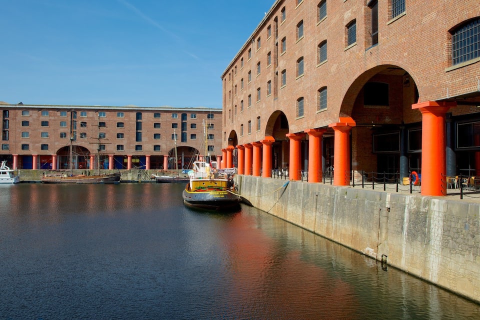 A river with a boat docked at a pier, a large brick building with arches, and a red-brick structure with columns.